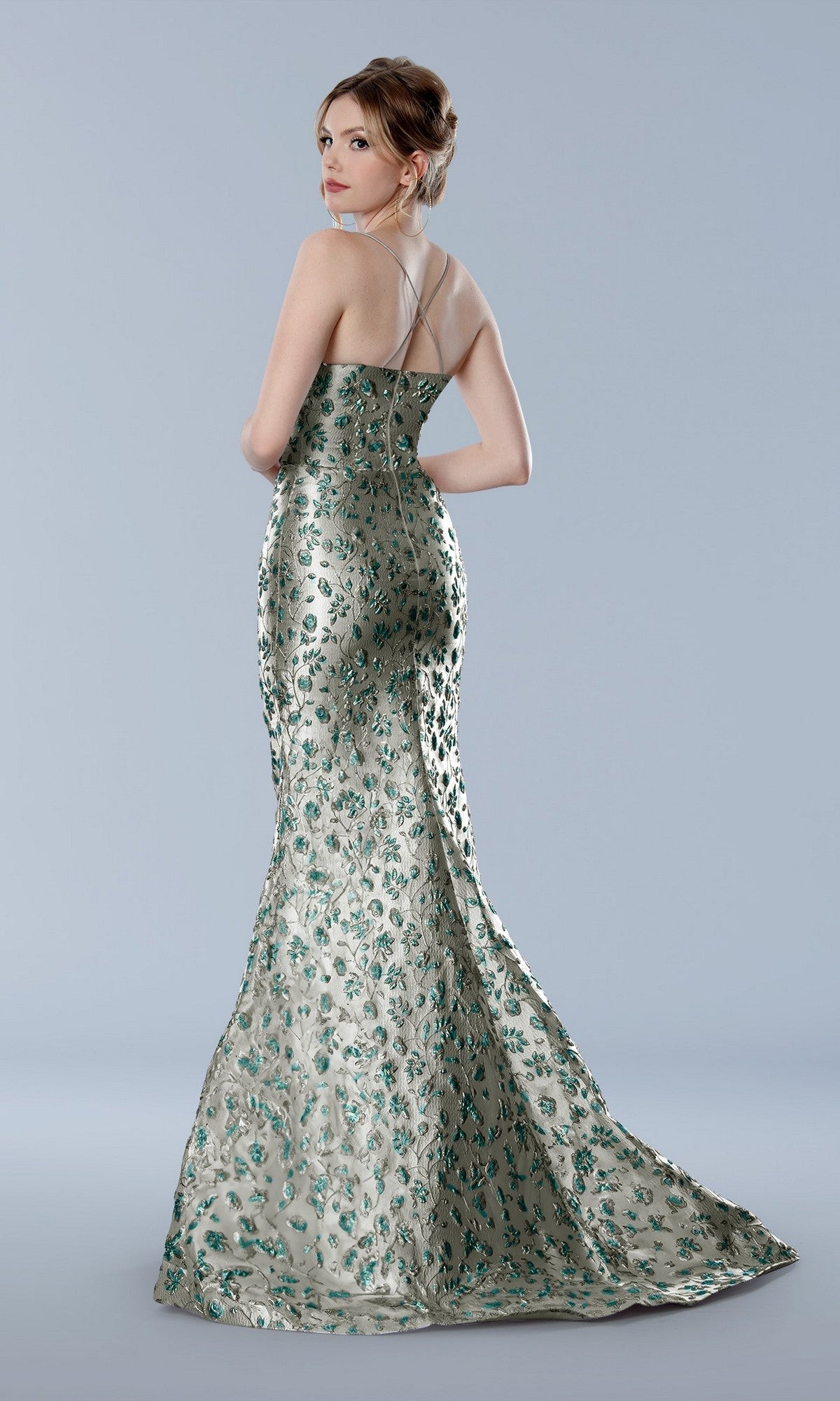 Woman in a sage Stella Couture 25328 long formal dress with thin straps, featuring green floral patterns, stands against a plain gray background.